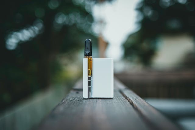 Cannabis vape cartridge and white box mod device on a wooden surface with blurred green background